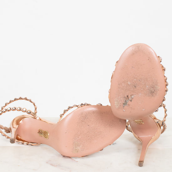 A pair of metallic rose gold high-heeled sandals with rose gold embellished straps and gold-tone hardware, photographed on a white marble surface against a white background.