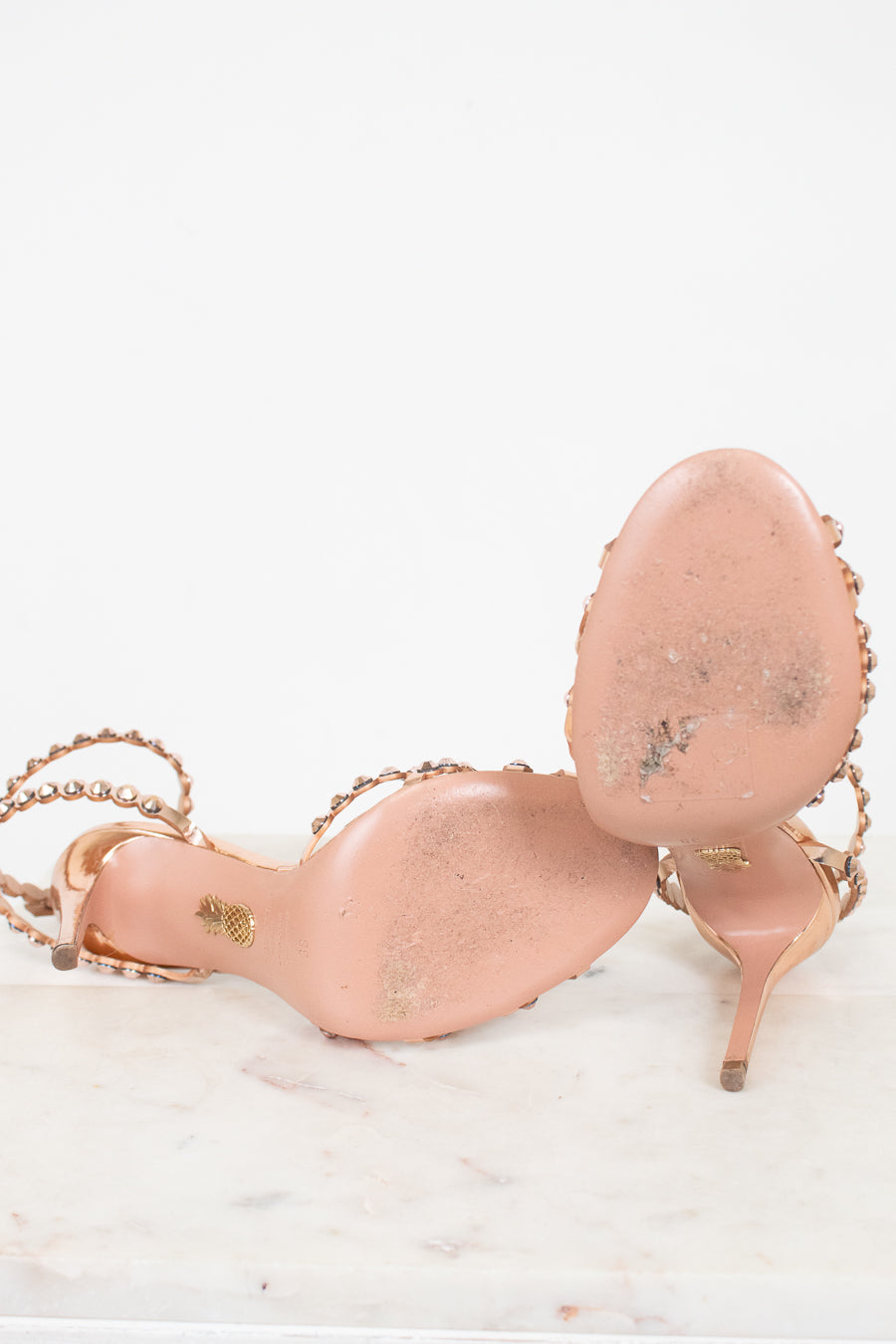 A pair of metallic rose gold high-heeled sandals with rose gold embellished straps and gold-tone hardware, photographed on a white marble surface against a white background.