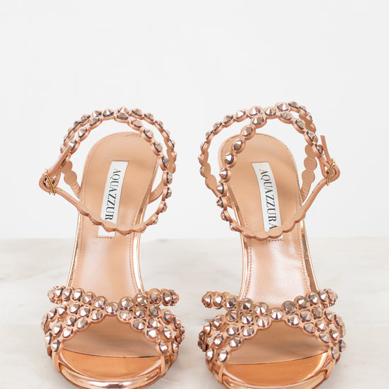 A pair of metallic rose gold heeled sandals with intricate rhinestone embellishments on the straps, displayed on a light marble surface against a white background. The brand name 'AQUAZZURA' is visible on the insole.