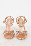A pair of metallic rose gold heeled sandals with intricate rhinestone embellishments on the straps, displayed on a light marble surface against a white background. The brand name 'AQUAZZURA' is visible on the insole.