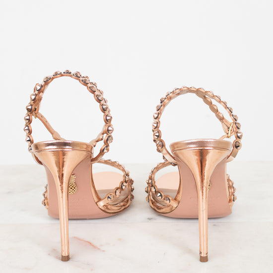 A pair of metallic rose gold high-heeled sandals with beaded straps, shown from the back on a light marble surface against a white background.