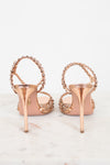 A pair of metallic rose gold high-heeled sandals with beaded straps, shown from the back on a light marble surface against a white background.