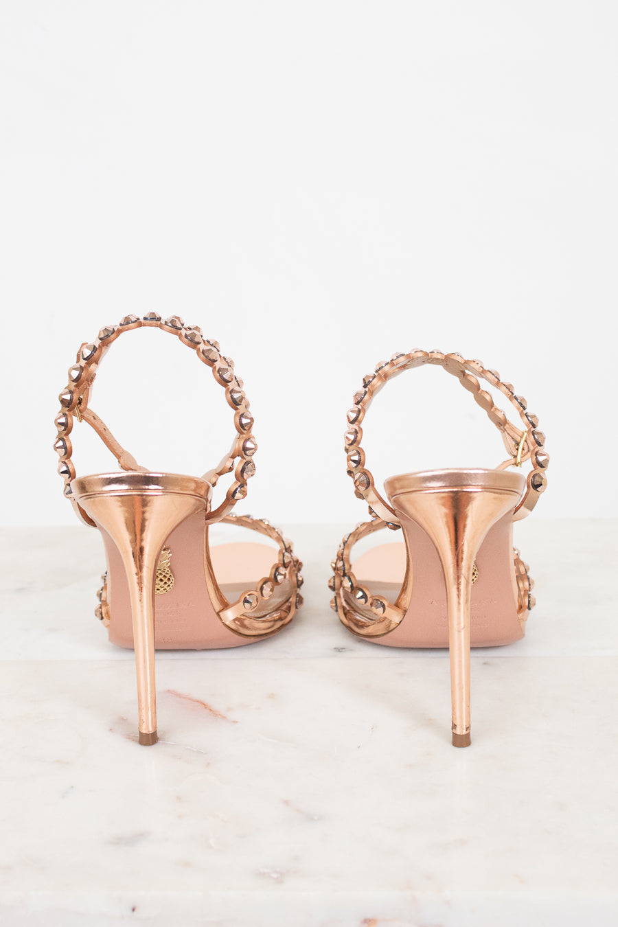 A pair of metallic rose gold high-heeled sandals with beaded straps, shown from the back on a light marble surface against a white background.