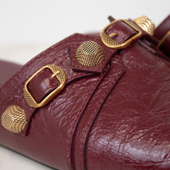 A close-up of a burgundy leather mule sandal featuring textured gold-tone metal buckles and studs, set against a light neutral background.