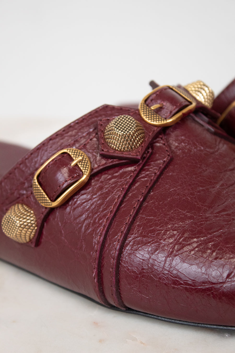 A close-up of a burgundy leather mule sandal featuring textured gold-tone metal buckles and studs, set against a light neutral background.