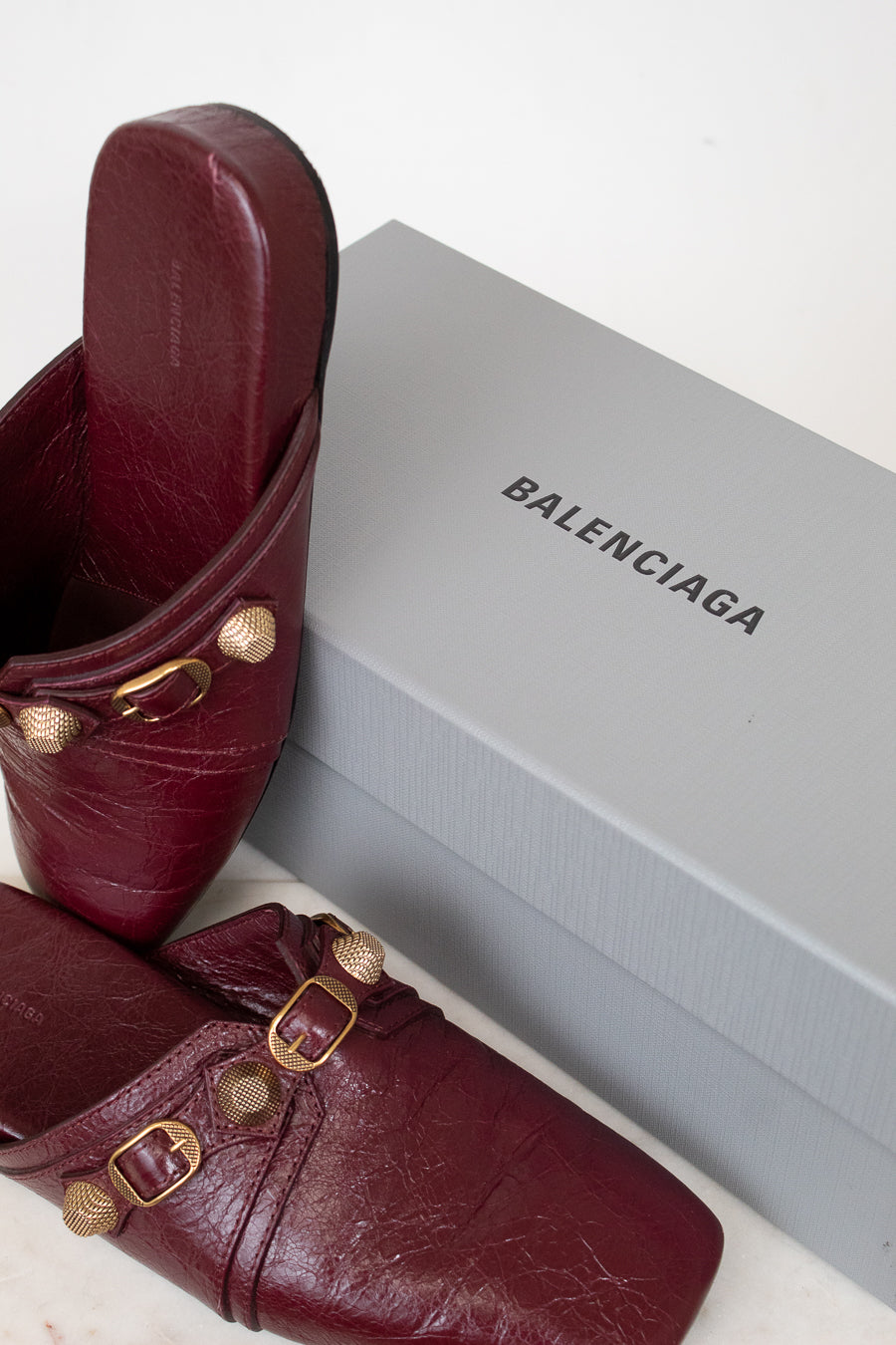 Burgundy mules with gold hardware set against a gray 'Balenciaga' branded box