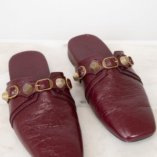 A pair of burgundy leather mule flats with gold-tone hardware, displayed on a light marble surface against a white background.