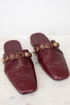 A pair of burgundy leather mule flats with gold-tone hardware, displayed on a light marble surface against a white background.