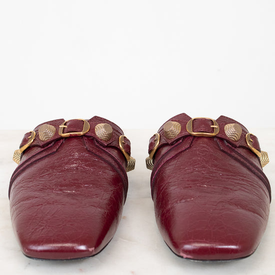 A pair of burgundy leather mule flats with gold-tone hardware and decorative studs, shown from the front on a white marble surface against a white background.