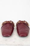 A pair of burgundy leather mule flats with gold-tone hardware and decorative studs, shown from the front on a white marble surface against a white background.