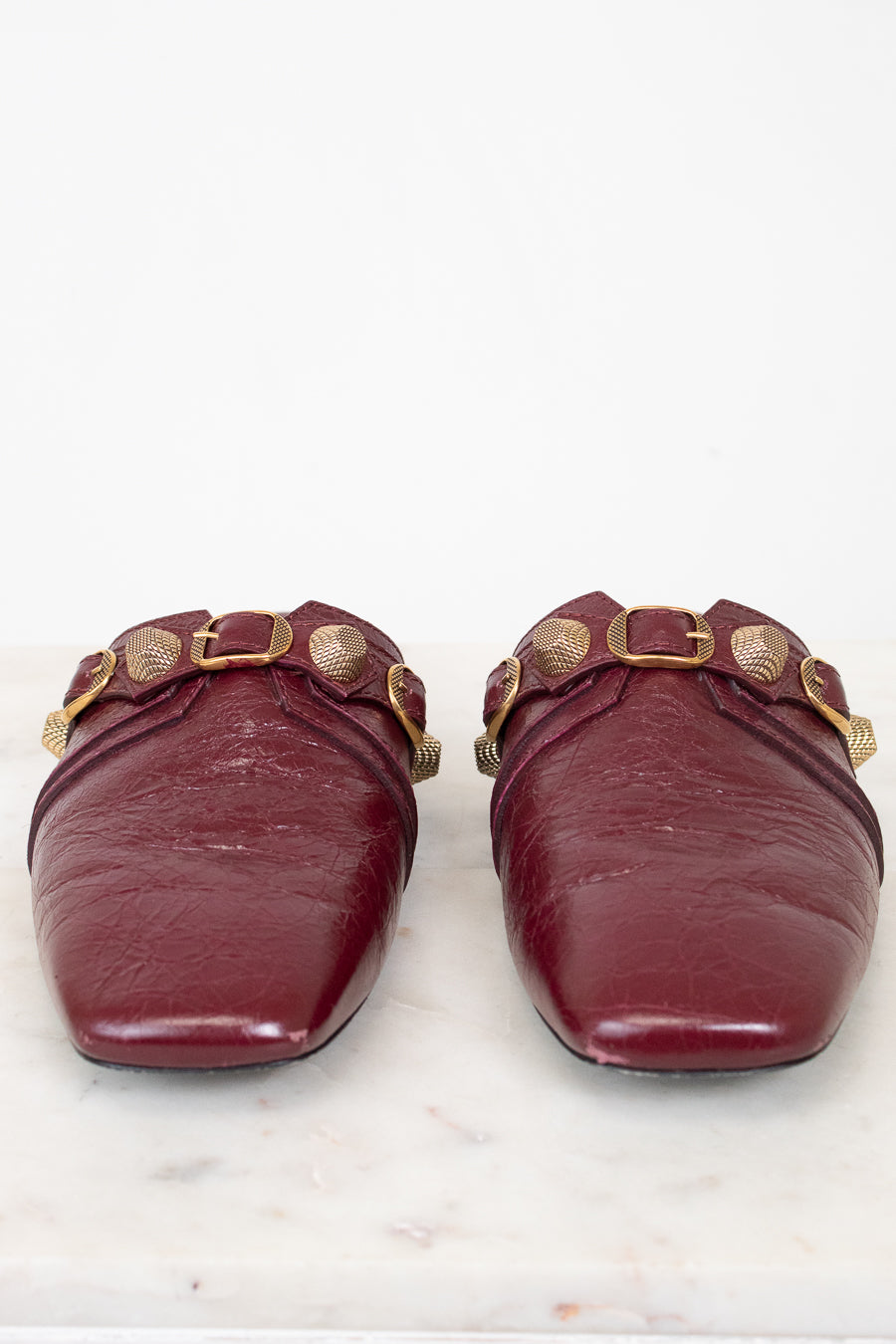 A pair of burgundy leather mule flats with gold-tone hardware and decorative studs, shown from the front on a white marble surface against a white background.