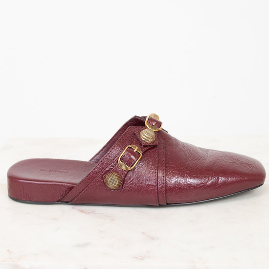 A single burgundy leather mule sandal with gold-tone buckle details, shown from a side profile on a white marble surface against a white background.