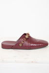 A single burgundy leather mule sandal with gold-tone buckle details, shown from a side profile on a white marble surface against a white background.