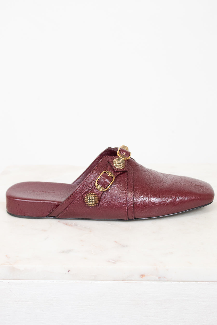 A single burgundy leather mule sandal with gold-tone buckle details, shown from a side profile on a white marble surface against a white background.
