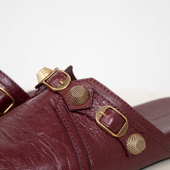 A close-up of a burgundy leather mule sandal featuring multiple gold-tone metal buckles and textured circular studs on the strap, set against a plain light background.