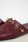 A close-up of a burgundy leather mule sandal featuring multiple gold-tone metal buckles and textured circular studs on the strap, set against a plain light background.