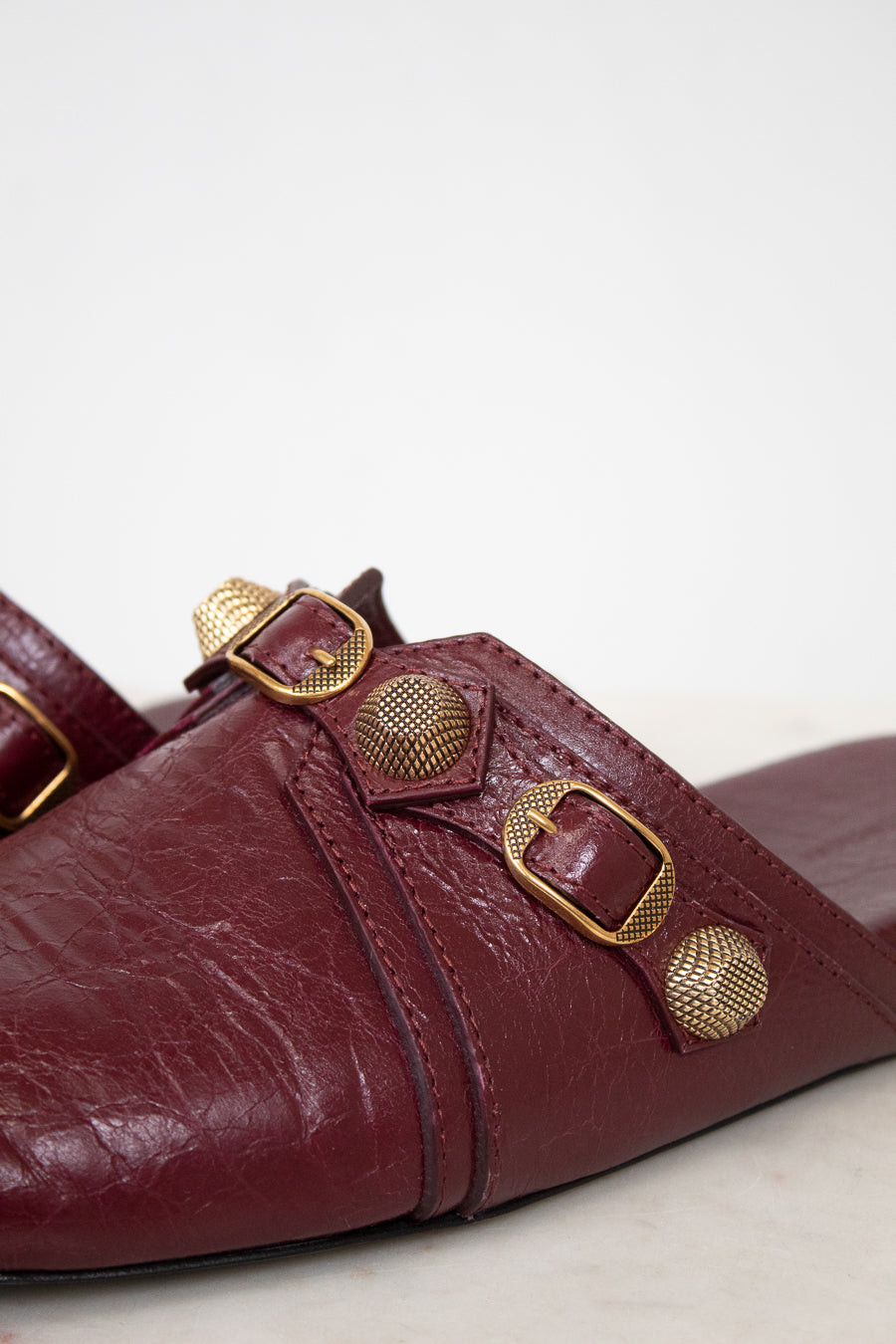 A close-up of a burgundy leather mule sandal featuring multiple gold-tone metal buckles and textured circular studs on the strap, set against a plain light background.