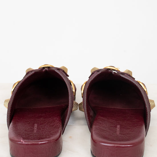 A pair of burgundy leather slide sandals with gold-tone chain and bow details, shown from a back-front view on a white marble surface against a white background.