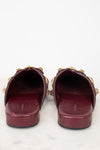 A pair of burgundy leather slide sandals with gold-tone chain and bow details, shown from a back-front view on a white marble surface against a white background.