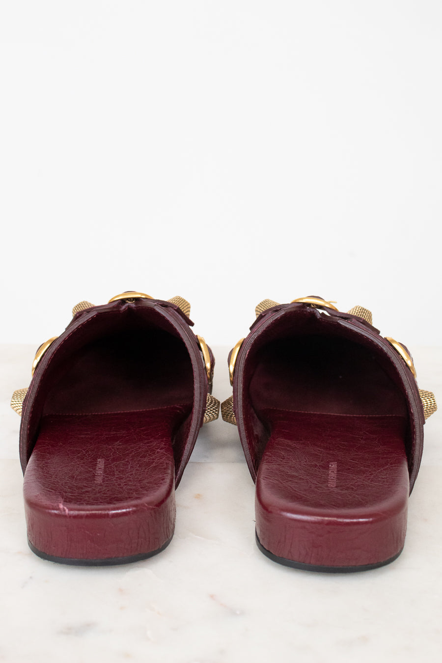 A pair of burgundy leather slide sandals with gold-tone chain and bow details, shown from a back-front view on a white marble surface against a white background.