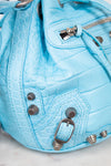 Close-up of a light blue textured handbag with metallic accents on a marble surface.