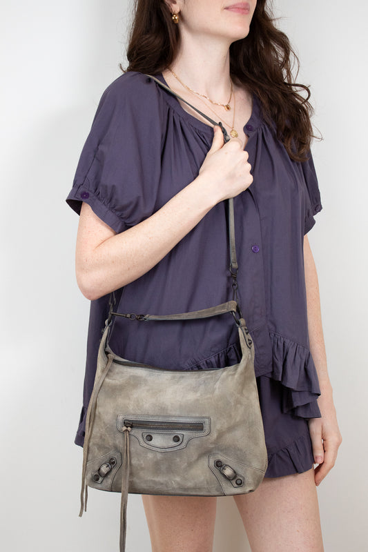 Woman wearing a purple blouse holding a gray shoulder bag against a white background