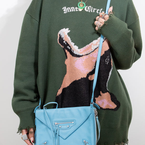 Person wearing a green sweater with a graphic design, holding a blue handbag against a plain background