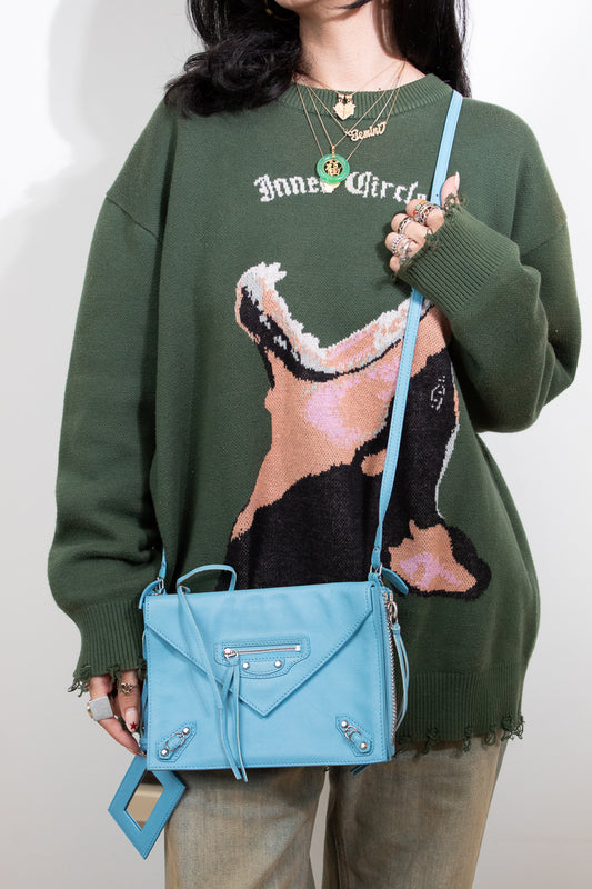 Person wearing a green sweater with a graphic design, holding a blue handbag against a plain background