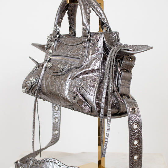 Front & side angle of silver metallic crinkled texture handbag with detailed rhinestone accents on a purse stand with a light background