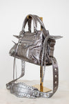 Front & side angle of silver metallic crinkled texture handbag with detailed rhinestone accents on a purse stand with a light background