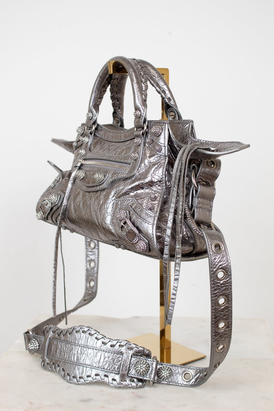 Front & side angle of silver metallic crinkled texture handbag with detailed rhinestone accents on a purse stand with a light background