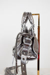 Side view of silver handbag with detailed straps on purse stand with a white background