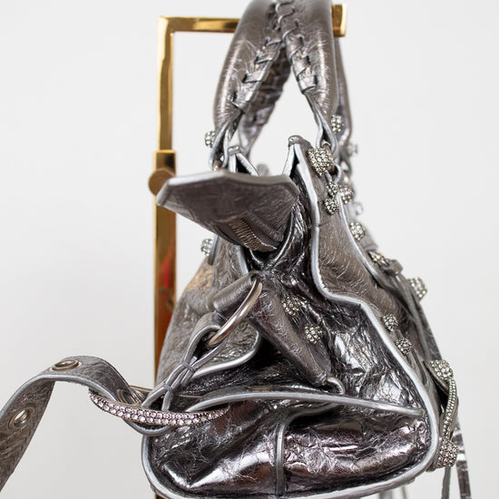 Side view of silver metallic handbag with decorative elements on a stand against a white background