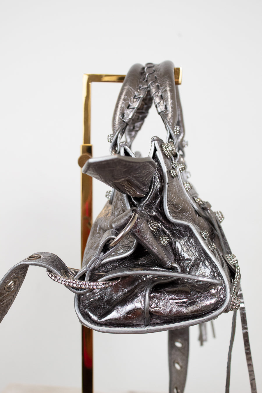 Side view of silver metallic handbag with decorative elements on a stand against a white background