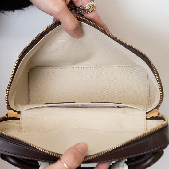 A close-up of a person's hands holding open a dark brown leather handbag to show its cream-colored interior lining and zippered compartments.