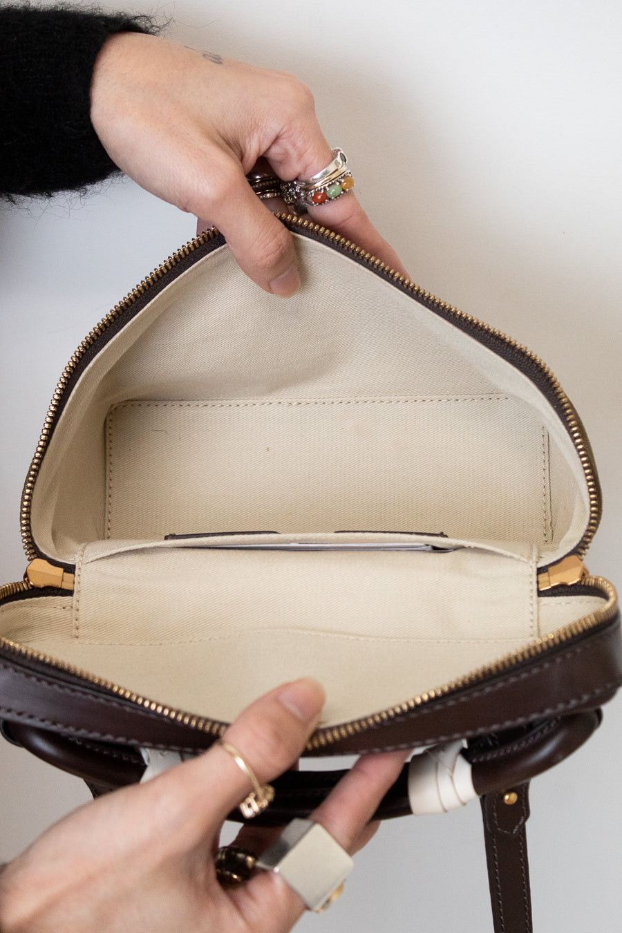 A close-up of a person's hands holding open a dark brown leather handbag to show its cream-colored interior lining and zippered compartments.