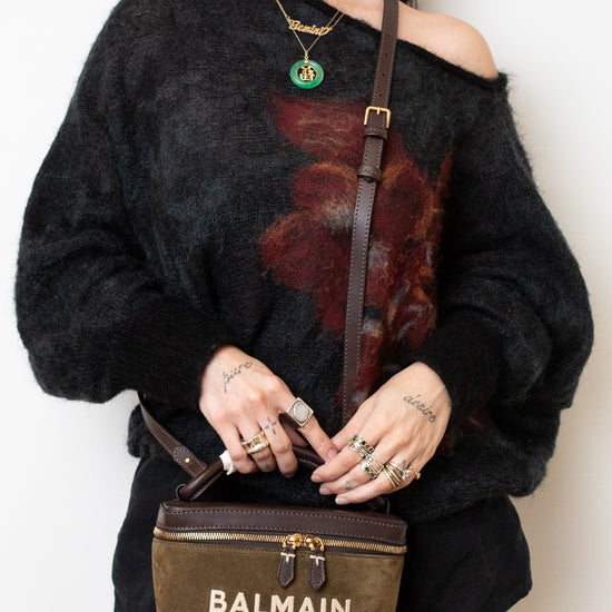 A woman wearing a black fuzzy off-the-shoulder sweater with a red floral print, holding a Balmain Paris brown suede crossbody bag with gold hardware.