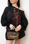 A woman wearing a black fuzzy off-the-shoulder sweater with a red floral print, holding a Balmain Paris brown suede crossbody bag with gold hardware.