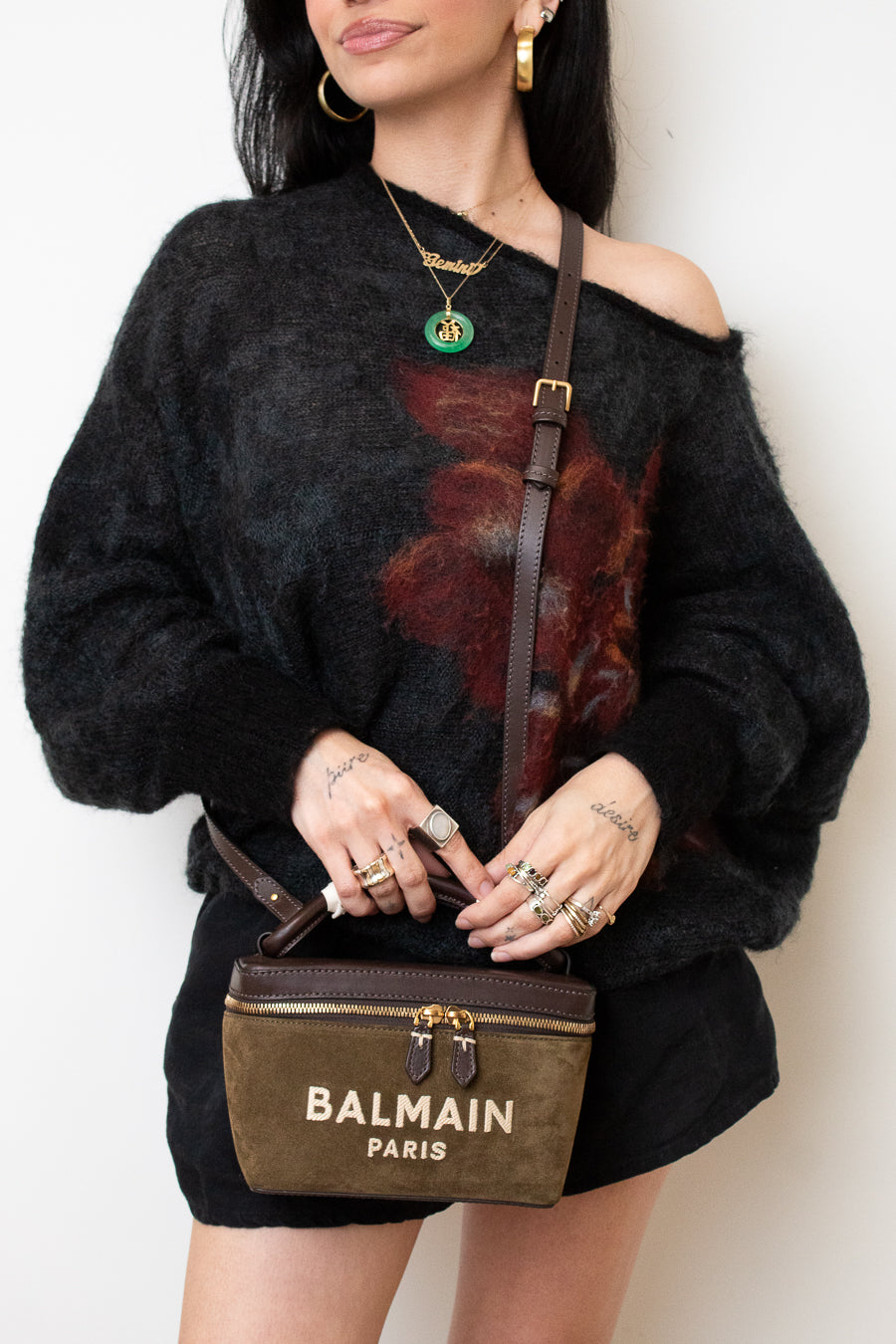A woman wearing a black fuzzy off-the-shoulder sweater with a red floral print, holding a Balmain Paris brown suede crossbody bag with gold hardware.