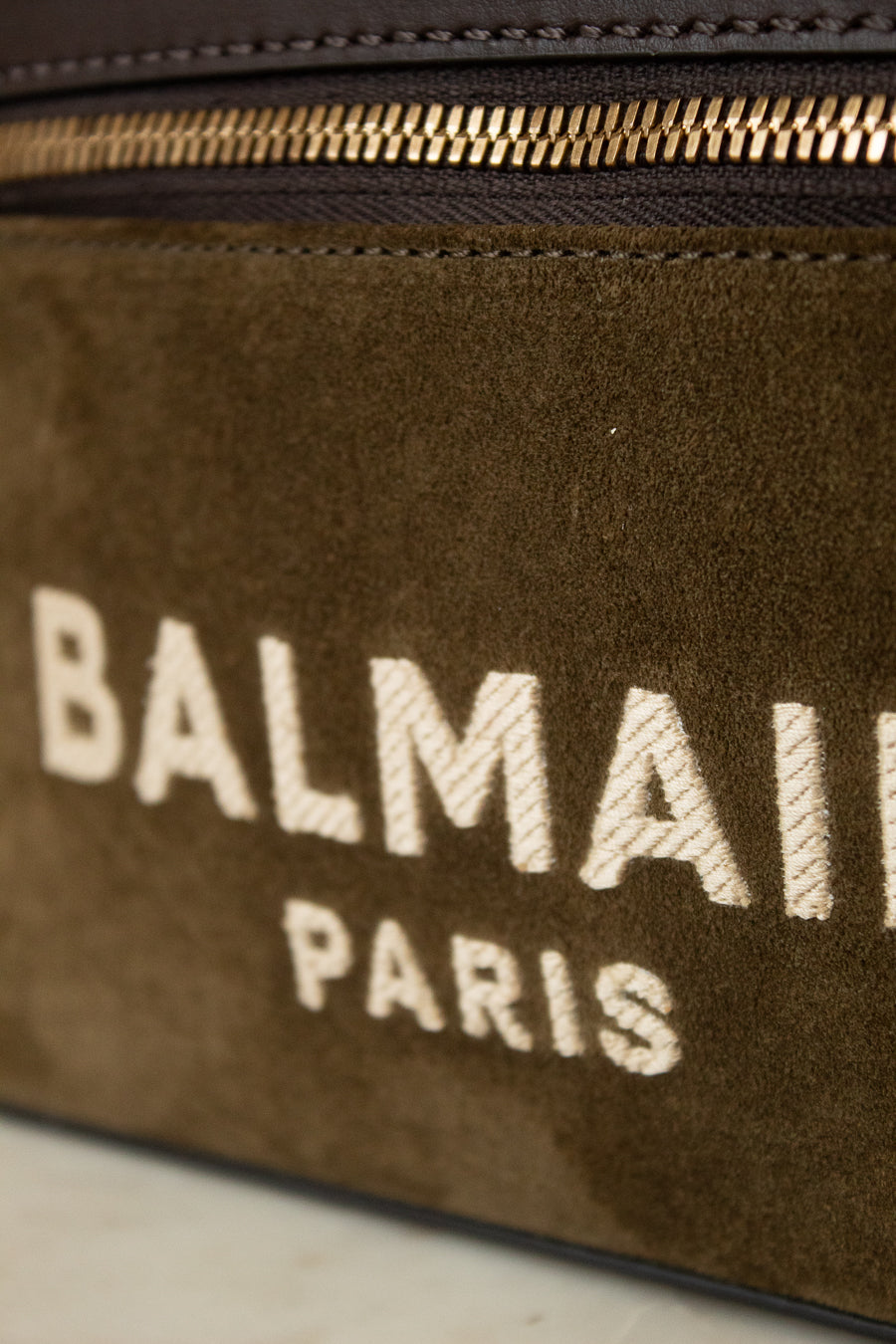 Close-up of a Balmain Paris suede bag in dark brown with a gold zipper and white embroidered logo text.