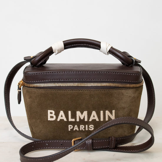 A Balmain Paris handbag in brown suede and dark brown leather, featuring a top handle wrapped in white ribbon, a gold zipper, and the 'BALMAIN PARIS' logo printed on the front, resting on a white marble surface against a white background.