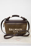 A Balmain Paris handbag in brown suede and dark brown leather, featuring a top handle wrapped in white ribbon, a gold zipper, and the 'BALMAIN PARIS' logo printed on the front, resting on a white marble surface against a white background.
