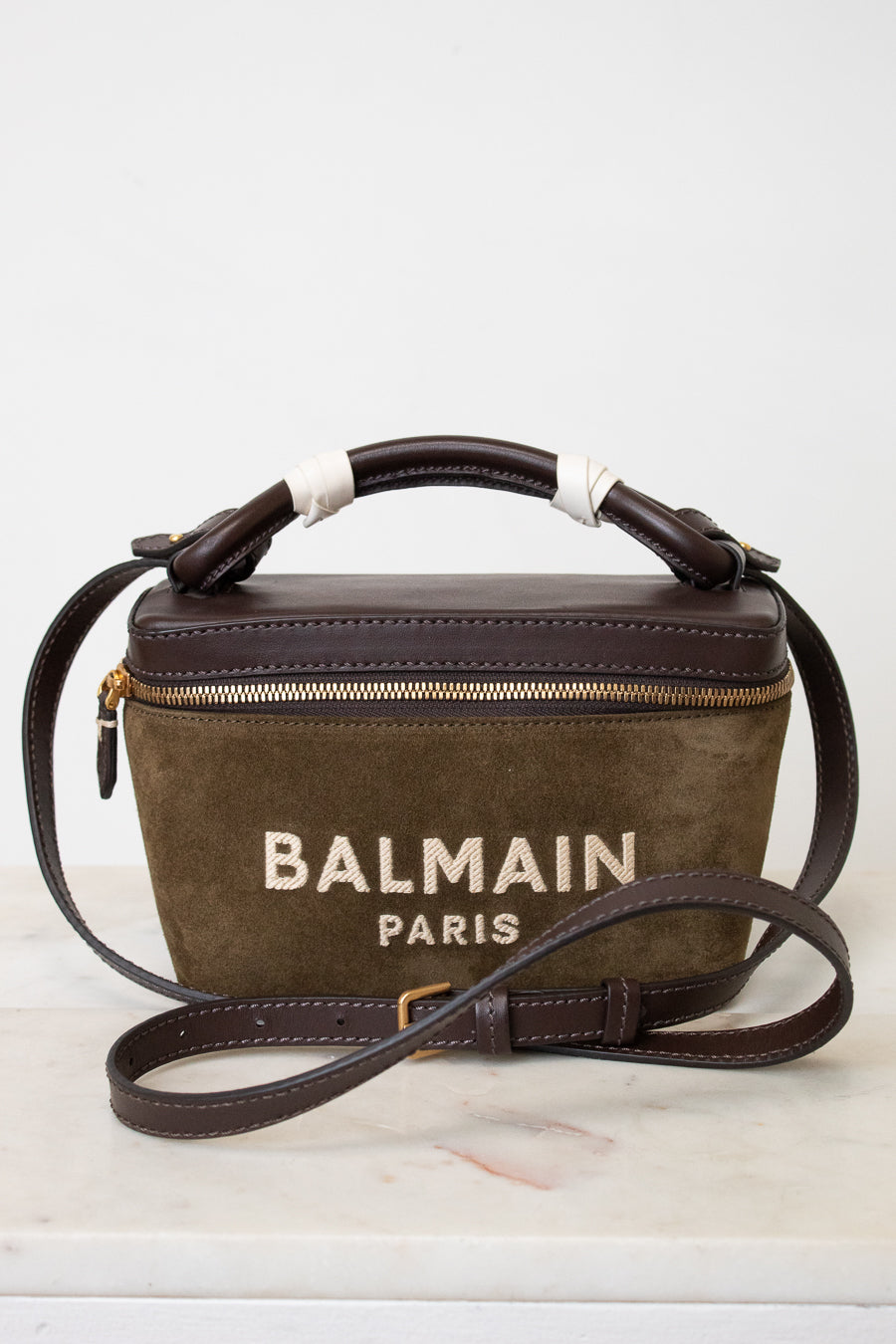 A Balmain Paris handbag in brown suede and dark brown leather, featuring a top handle wrapped in white ribbon, a gold zipper, and the 'BALMAIN PARIS' logo printed on the front, resting on a white marble surface against a white background.