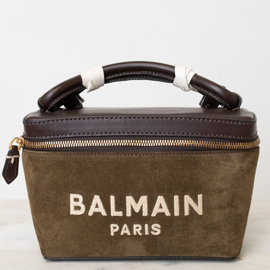 A Balmain Paris handbag in olive green suede with dark brown leather trim and handle, featuring gold hardware and white embroidered branding on the front, set against a light background.