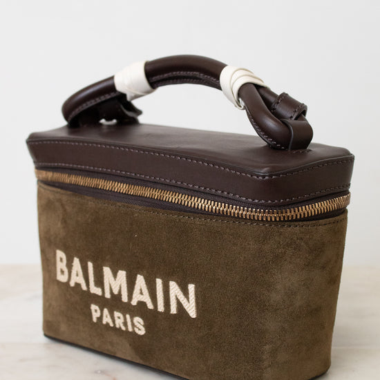 A Balmain Paris handbag in brown suede and dark brown leather, featuring a top handle wrapped in white bands and a gold zipper, set against a light background.