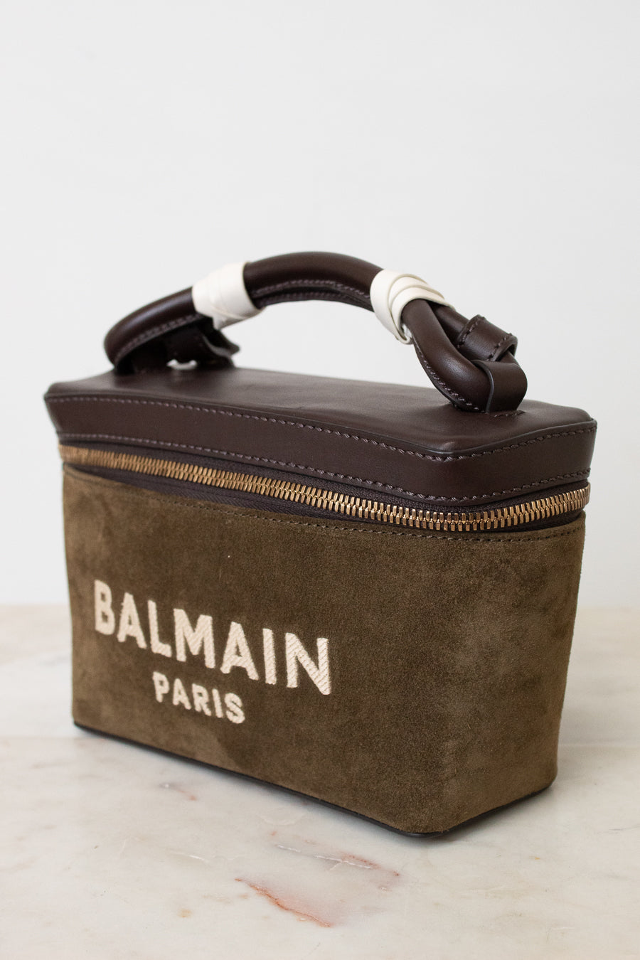 A Balmain Paris handbag in brown suede and dark brown leather, featuring a top handle wrapped in white bands and a gold zipper, set against a light background.