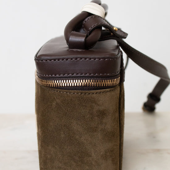Side profile of a small bag showing brown leather and brown suede. Resting on a marble top and white background.