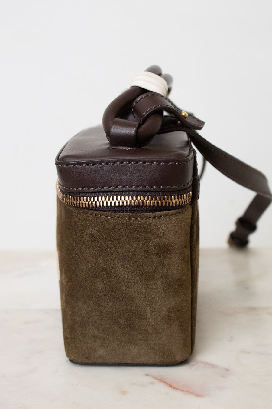 Side profile of a small bag showing brown leather and brown suede. Resting on a marble top and white background.