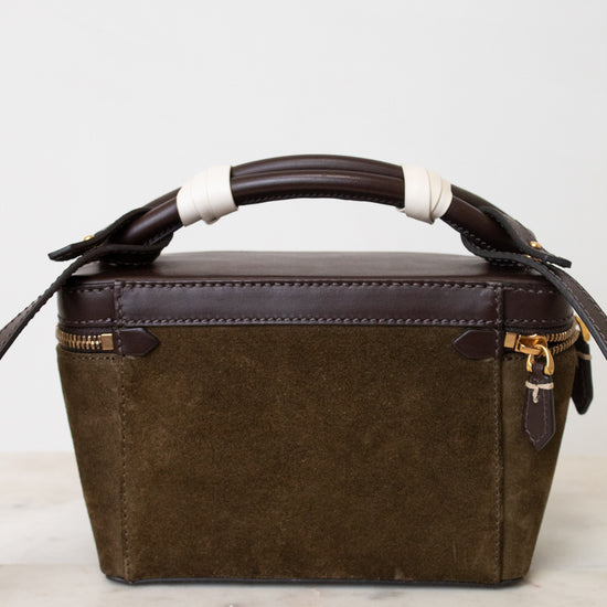 A structured rectangular handbag in dark brown leather and olive green suede, featuring a wrapped bamboo handle and gold-tone hardware, set against a clean off-white background.