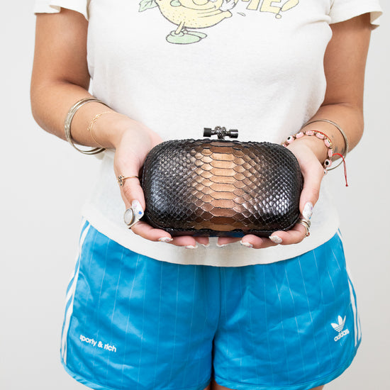 Person holding a black snake-textured clutch bag wearing a white t-shirt with a graphic and blue shorts.